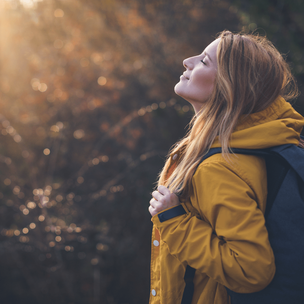 woman in a rain coat and hiking backpack looking up at the sun in the forest